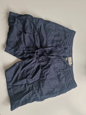 Mörkblå linneshorts med resårmidja - Snygga mörkblå shorts i linne med avslappnad passform och resår i midjan. Shortsen har sidofickor och bakfickor, perfekta för varma dagar. Passar dig som gillar en enkel och stilren look.