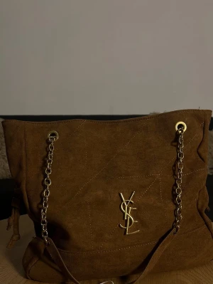 Brun  handväska från YSL - Snygg brun handväska i  från YSL med guldiga kedjehandtag och ikonisk YSL-logga framtill. Väskan har en mjuk, rymlig form och detaljer i guldmetall. Perfekt för dig som gillar lyxiga accessoarer med trendig känsla.