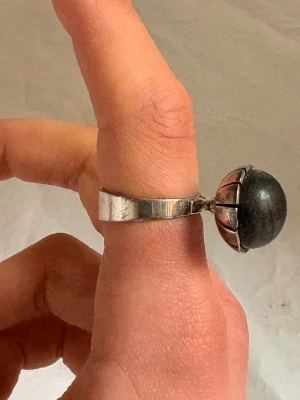 Silverring med stor svart sten - Cool ring i silver med en stor, rund svart sten som sitter i en kloliknande fattning. Ringen har en bred, slät ringskena och den svarta stenen ger ett unikt och iögonfallande intryck. Perfekt för dig som gillar statement-smycken.