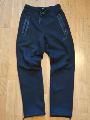 Svarta baggy Nike tech byxor - Svarta mjukisbyxor från Nike med dragsko i midjan och två dragkedjeförsedda fickor framtill. Byxorna har en relaxed passform och är tillverkade i mjukt material som passar perfekt för chill eller träning. Nike-logga på vänster sida.