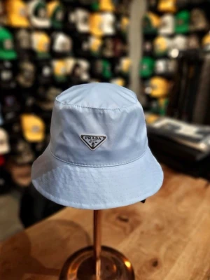 Ljusblå bucket hat från Prada - Stilren ljusblå bucket hat från Prada med klassisk siluett och ikonisk metallogo framtill. Hatten är tillverkad i ett lätt och bekvämt material som passar perfekt för en trendig look.