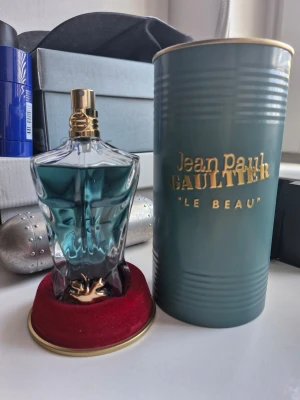 Jean Paul Gaultier Le Beau parfym - Unik parfymflaska från Jean Paul Gaultier Le Beau med blågrön vätska och en kroppsinspirerad glasdesign. Flaskan har gulddetaljer och kommer med en elegant grön metalltub med guldfärgad text. Perfekt för dig som gillar snygg design och ikoniska dofter. Hör av er vid frågor och priset är ej hugget i sten.