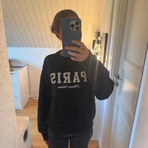 Svart sweatshirt med Paris-tryck - Svart sweatshirt från H&M i storlek XS med stort Paris-tryck i vitt framtill. Tröjan har rund hals, ribbade muddar vid ärmslut och nederkant samt mjukt, fleeceliknande foder på insidan. 