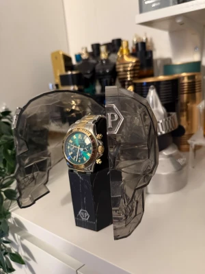 Philipp Plein klocka guld och silver - Snygg Philipp Plein klocka i guld och silver med grön urtavla och detaljer i guld. Klockan har en robust design och levereras i en unik förpackning formad som ett dödskallehuvud. Perfekt accessoar för dig som vill sticka ut. Alla orginal grejer följer med så som box, kartong, tagg osv. Kan tänka mig byta mot annan klocka, parfymer osv.