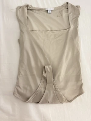 Beige body med fyrkantig ringning - Snygg beige body från Amisu med fyrkantig ringning och ärmlös design. Tillverkad i ett mjukt och stretchigt material som sitter skönt mot kroppen. Perfekt att styla med jeans eller kjol för en trendig look.
