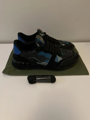 Valentino Garavani camo sneakers - Snygga sneakers från Valentino Garavani med svart bas och blått, grått och vitt camomönster. Skorna har svarta snören, detaljer i läder och textil samt en cool nitar-detalj på sulan. Perfekta för dig som vill sticka ut med exklusiv streetstyle.