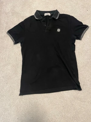 Svart pikétröja från Stone Island - Svart pikétröja från Stone Island med klassisk krage och knappar framtill. Tröjan har vita ränder på krage och ärmslut samt en broderad logotyp på bröstet. Perfekt för en stilren och avslappnad look.