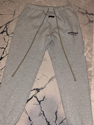 Grå Essentials mjukisbyxor - Snygga grå mjukisbyxor från Essentials Fear of God med elastisk midja och långa ben. Byxorna har långa snören framtill och tryckt logga på vänster lår. Perfekta för en chill och avslappnad stil. Pris går att diskutera.