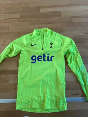 Neongul Tottenham träningströja Nike - Säljer en neongul långärmad träningströja från Nike med Tottenham Hotspur-logga och sponsortryck 'getir' på bröstet. Tröjan har halv dragkedja framtill och är perfekt för fotbollsträning eller uppvärmning. Dri-Fit