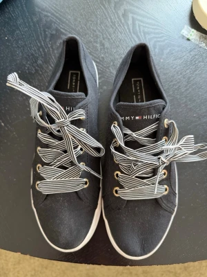 Svarta Tommy Hilfiger sneakers - Svarta låga sneakers från Tommy Hilfiger med vita och svarta randiga skosnören och guldfärgade öljetter. Snygg logga på plösen och klassisk vit sula. Perfekta för en stilren och avslappnad look. Aldrig använda. 