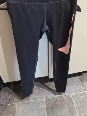Svarta leggings från Nike med rosa detalj - Snygga svarta leggings från Nike i storlek XS med en cool rosa swoosh-detalj längs ena benet. Perfekta för träning eller chill, med stretchigt material som sitter skönt och följer kroppen. Klassisk Nike-kvalitet och stilren design.