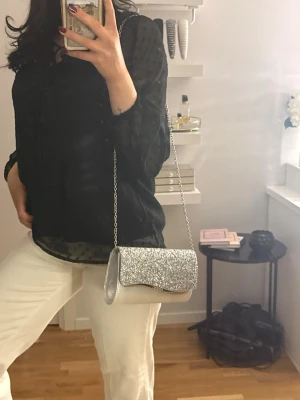 Silverglittrig kuvertväska med kedja - Säljer en snygg kuvertväska i silver med glittrig yta och kedjerem. Väskan har en elegant, avlång form och stängs med lock. Perfekt för dig som vill sticka ut och addera lite extra bling till din outfit.
