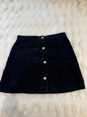 Svart manchesterkjol med knappar - Snygg svart kortkjol i manchester med raka linjer och silverfärgade knappar framtill. Kjolen har en klassisk midja och är perfekt för dig som gillar en stilren och trendig look.