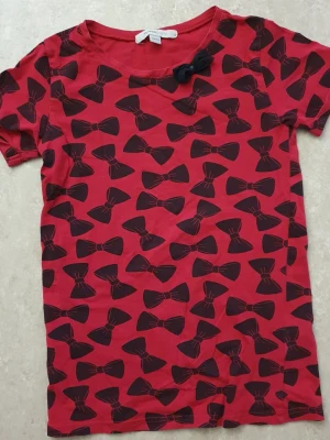 T-shirt - Cool röd t-shirt från Mywear Young med svarta rosetter över hela tröjan och en söt svart rosett vid halsen. T-shirten har korta ärmar och är gjord i mjuk bomull, perfekt för en trendig vardagslook.