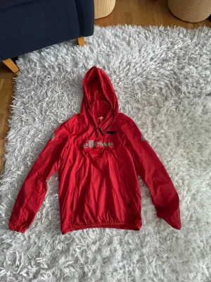 Röd vindjacka från Ellesse med huva - Snygg röd vindjacka från Ellesse med stor logga på bröstet. Jackan har huva med dragsko, dragkedja vid halsen och elastisk nederkant. Perfekt för blåsiga dagar och enkel att matcha med streetwear. Materialet är lätt och vattenavvisande.
