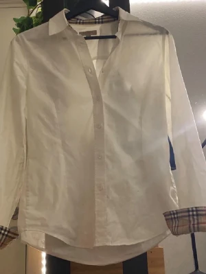 Vit burberry skjorta - Klassisk vit skjorta från Burberry med ikoniska rutiga detaljer på insidan av kragen och ärmsluten. Skjortan har knappar framtill och lång ärm, perfekt för en stilren look.
