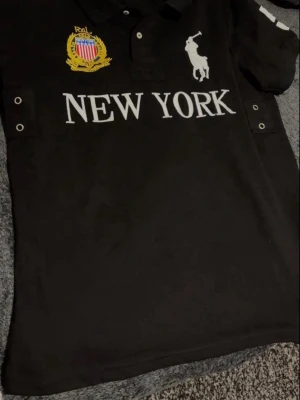 Svart pikétröja Polo Ralph Lauren - Svart pikétröja från Polo Ralph Lauren med stor vit logga, broderad gul emblem och texten NEW YORK framtill. Klassisk krage med knappar och metallöljetter på sidorna. Perfekt för en sportig och stilren look.