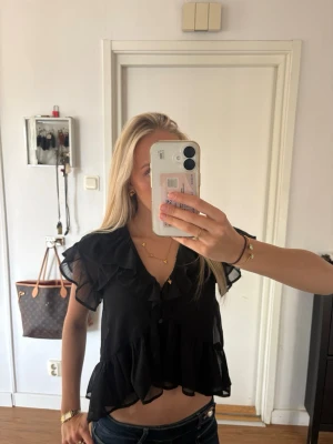 Svart volangblus med knappar - Snygg svart blus med volanger och knappar framtill. Blusen har korta, luftiga ärmar i transparent material och en v-ringad halsringning. Perfekt för en trendig och chic look. Materialet känns lätt och flowy.