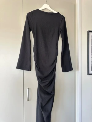Svart långklänning  - Svart långklänning från Boohoo med lång ärm och rund halsringning. Klänningen har en tight passform och snygg rynkdetalj längs sidan som ger en cool siluett. Tillverkad i ett stretchigt syntetmaterial som sitter bekvämt på kroppen. Endast använd 1 gång!