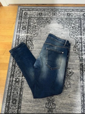 Blå slitna skinny jeans med detaljer - Säljer ett par blå skinny jeans med slitningar och patchade detaljer framtill. Jeansen har klassisk femficksdesign och en snygg tvätt med ljusare partier. Perfekta för en trendig och avslappnad look.