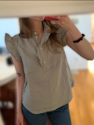 Khakifärgad blus med volangkrage - Supersöt khakifärgad blus med korta volangärmar och rynkad struktur i 100% bomullsmuslin. Blusen har en liten ståkrage med volangdetaljer och knappar framtill. Perfekt till jeans eller kjol för en trendig look. Strl S, passar även XS. Aldrig använd så i utmärkt skick, däremot är prislappen ej kvar.