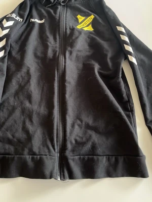 Svart Hummel träningsjacka BK Öksten - Svart träningsjacka med dragkedja från Hummel, med BK Öksten-logga på bröstet. Jackan har vita detaljer på ärmarna och huva. Perfekt för träning eller uppvärmning. Materialet är troligtvis polyesterblandning.