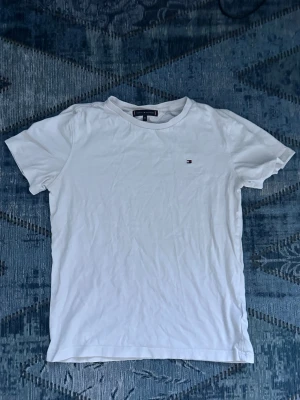 Vit t-shirt från Tommy Hilfiger - Klassisk vit t-shirt från Tommy Hilfiger med liten broderad logga på bröstet. T-shirten har rund halsringning och korta ärmar. Tillverkad i mjuk bomull för en skön känsla och enkel stil.