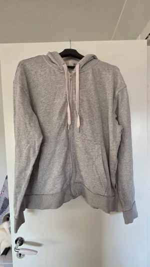 Grå zip-hoodie från H&M XL - En klassisk grå hoodie med dragkedja från H&M. Hoodien har huva med vita dragsnören, två fickor framtill och ribbade muddar vid ärmslut och nederkant. Perfekt för en chill och avslappnad stil.