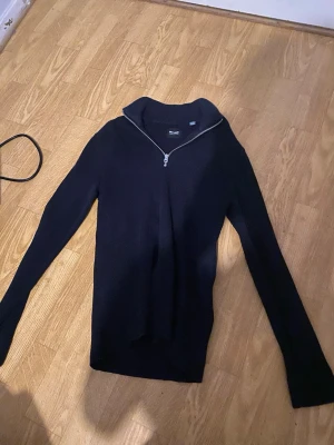 Svart stickad tröja med halv dragkedja - Svart stickad tröja från Blend med lång ärm och halv dragkedja vid halsen. Tröjan har en hög krage och är tillverkad i ett mjukt, ribbstickat material. Perfekt för lager-på-lager och enkel att matcha med andra plagg.från only sons 