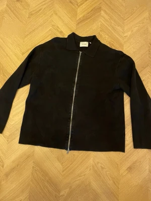 Svart tröja  från Sandö  med dragkedja - Stilren svart tröja från Sandö med rak passform och klassisk krage. Jackan har en hel dragkedja framtill och är tillverkad i ett mjukt bomullstyg. Perfekt att slänga på sig över en t-shirt för en clean look.