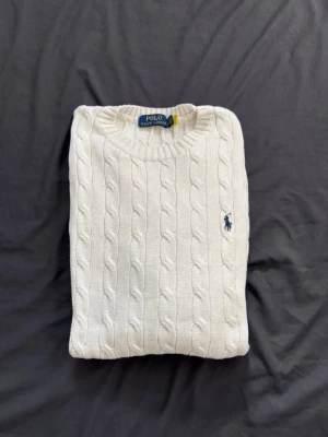 Vit kabelstickad tröja Polo Ralph Lauren - Vit kabelstickad tröja från Polo Ralph Lauren med rund halsringning och klassisk blå logga broderad på bröstet. Tröjan har ribbade muddar och är tillverkad i mjukt material som passar perfekt till lager-på-lager-stil.