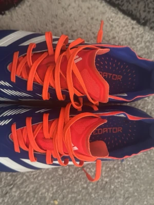 Adidas Predator blå fotbollsskor - Snygga Adidas Predator fotbollsskor i blått med vita detaljer och orange snörning. Skorna har en syntetisk ovandel och en orange sula med fasta dobbar för bra grepp på planen. Perfekta för dig som vill sticka ut på matchen!