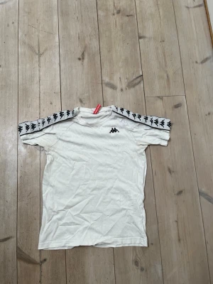 Vit Kappa t-shirt med logoband - Vit t-shirt från Kappa med klassiska svarta logoband längs axlarna denna modell är väldigt ovanlig i denna storlek i Sverige köptes i Italien 