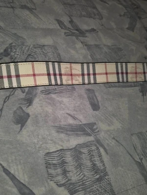 Beige rutigt bälte med B-spänne - Stilrent bälte i beige med klassiskt rutigt mönster i svart, vitt och rött. Bältet har ett stort guldfärgat B-spänne som ger en snygg detalj. Perfekt accessoar för att lyfta din outfit.
