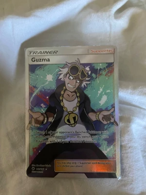Pokémonkort Guzma Trainer Supporter - Säljer ett Pokémonkort av typen Trainer Supporter med karaktären Guzma. Kortet har färgglad bakgrund och tydlig text på framsidan. Baksidan har klassiskt Pokémon-motiv med Pokéboll. Perfekt för samlare eller Pokémon-fans.