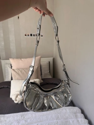 Silvrig axelremsväska i metallic - Trendig handväska i metallic-silver med justerbar axelrem och dragkedja upptill. Väskan har en rundad form och veckade detaljer framtill som ger en cool look. Perfekt accessoar för att lyfta din outfit.