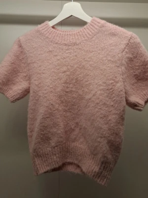 Ljusrosa stickad t-shirt från Bik Bok - Supersöt ljusrosa stickad t-shirt från Bik Bok i fluffigt material. Tröjan har korta ärmar, bred ribbad halsringning och ribbade muddar nedtill. Perfekt för dig som gillar mjuka och mysiga plagg med en feminin touch.