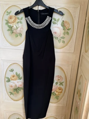 Svart klänning med pärldetaljer Zara - Elegant svart klänning från Zara Woman med pärlbroderad halsringning. Klänningen har ärmlös design, figurnära passform och stilrena veckdetaljer framtill. 