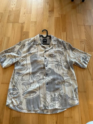 Beige och grå skjorta från Only & Sons med mönster - Kortärmad skjorta från Only & Sons i beige och grått med ett unikt abstrakt mönster av ansikten och former. Skjortan har knappar framtill och en avslappnad passform.