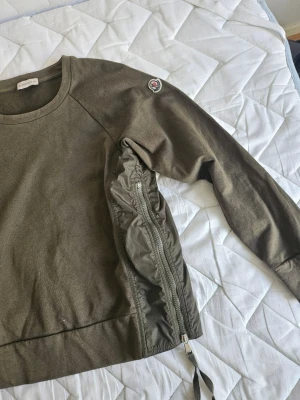 Olivgrön sweatshirt med dragkedja - Cool äktal olivgrön sweatshirt med rund hals och lång ärm. Unik detalj med dragkedja och snörning längs sidan samt patch på ena ärmen. Tillverkad i mjukt bomullsmaterial med inslag av nylon vid dragkedjan. Perfekt för dig som gillar streetwear och vill sticka ut.