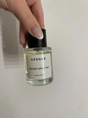 ESSNCE Drunk Apple Pie parfym - ESSNCE Drunk Apple Pie Eau de Parfum i en rund glasflaska med svart lock. Doften har en fräsch och söt ton av äppelpaj. Flaskan har en minimalistisk vit etikett med svart text och känns modern och stilren.