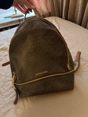 Brun ryggsäck från Michael Kors - Snygg brun ryggsäck från Michael Kors med klassiskt MK-mönster över hela väskan. Väskan har guldfärgade dragkedjor, justerbara axelband i brunt läder. 