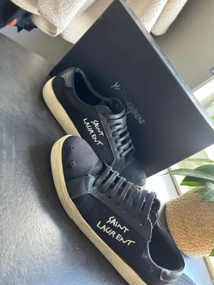 Svarta Saint Laurent sneakers - Svarta sneakers från Saint Laurent med vit textlogga på sidan. Skorna har en klassisk låg siluett, svarta skosnören och vit sula. Materialet är en mix av canvas och läderdetaljer vilket ger en stilren och trendig look.