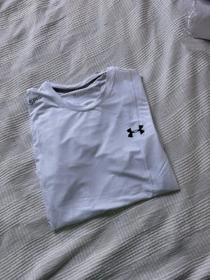 Vit träningströja från Under Armour - Snygg vit t-shirt från Under Armour med svart logga på bröstet och tryck på ärm och nederkant. Tillverkad i ett lätt och snabbtorkande funktionsmaterial som passar perfekt till träning och sportaktiviteter. Klassisk rund hals och korta ärmar. Storlek L men sitter som M