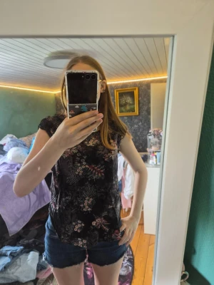 Blommig topp från ONLY - Snygg svart topp från ONLY med blommigt mönster i rosa och vitt. Modellen har korta ärmar och en avslappnad passform, perfekt till jeans eller shorts. Tunt och mjukt material som känns skönt mot huden.