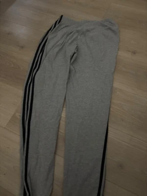 Grå Adidas mjukisbyxor med svarta ränder - Grå mjukisbyxor från Adidas med klassiska svarta ränder längs sidorna och elastisk midja. Perfekta för chill eller träning, med en clean och sportig look. Adidas-logga på ena benet. Mjukt och bekvämt material.