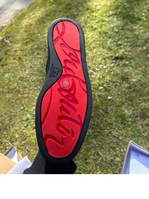 Christian Louboutin svarta sneakers - Svarta sneakers från Christian Louboutin med klassisk röd sula och svarta nitar på tån. Skon är i mocka och läder, har svarta skosnören och låg profil. Perfekt för dig som vill sticka ut med en edgy och lyxig stil.