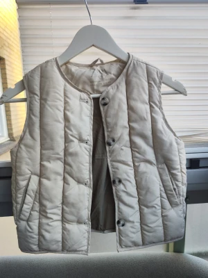 Beige dunväst från Zara barn storlek 110 - Snygg beige dunväst från Zara för barn, med raka sömmar och knäppning framtill. Västen har två fickor och är lätt vadderad för extra värme. Perfekt att slänga över en hoodie eller tröja när det är kyligt ute. Storlek: 110. Aldrig används innan, helt ny väst!