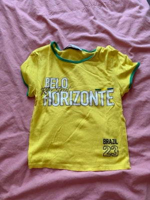 Gul t-shirt från Berskha - Gul t-shirt med gröna detaljer och texten 'Belo Horizonte' samt 'Brazil 23' framtill. T-shirten har korta ärmar och en avslappnad passform. Perfekt för dig som gillar sportig stil och Brasilien-vibbar.