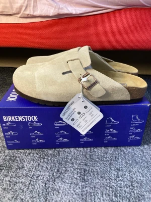 Beige Birkenstock Boston tofflor - Säljer ett par klassiska Birkenstock Boston tofflor i beige mocka med justerbart spänne i metall. Fotbädden är mjuk och anatomiskt utformad för extra komfort. Perfekta för en avslappnad och stilren look. Levereras i originalkartong.